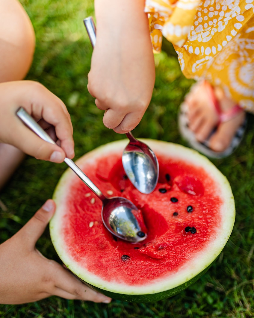 Two caucasian children sitting on grass spooning out watermelon from the fruit.