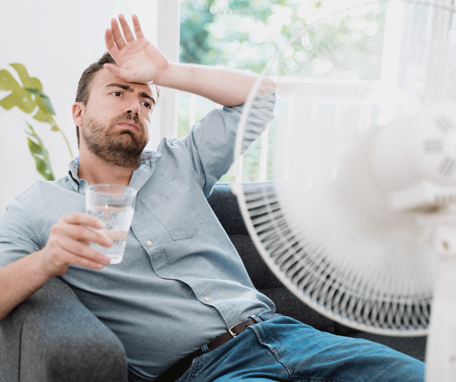 A man sitting on a couch in front of a fan drinking a glass of water to cool down. 