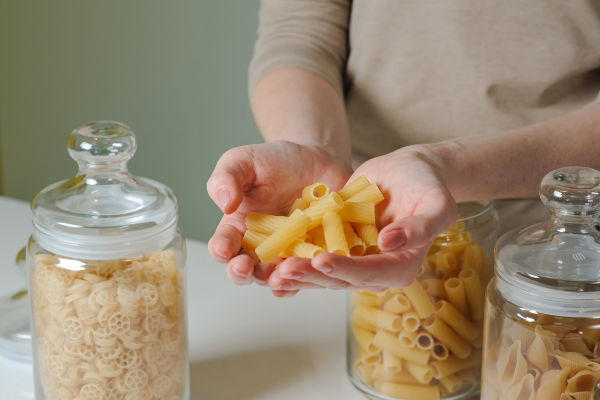 Woman with a handful of pasta over three glass jars with raw pasta.